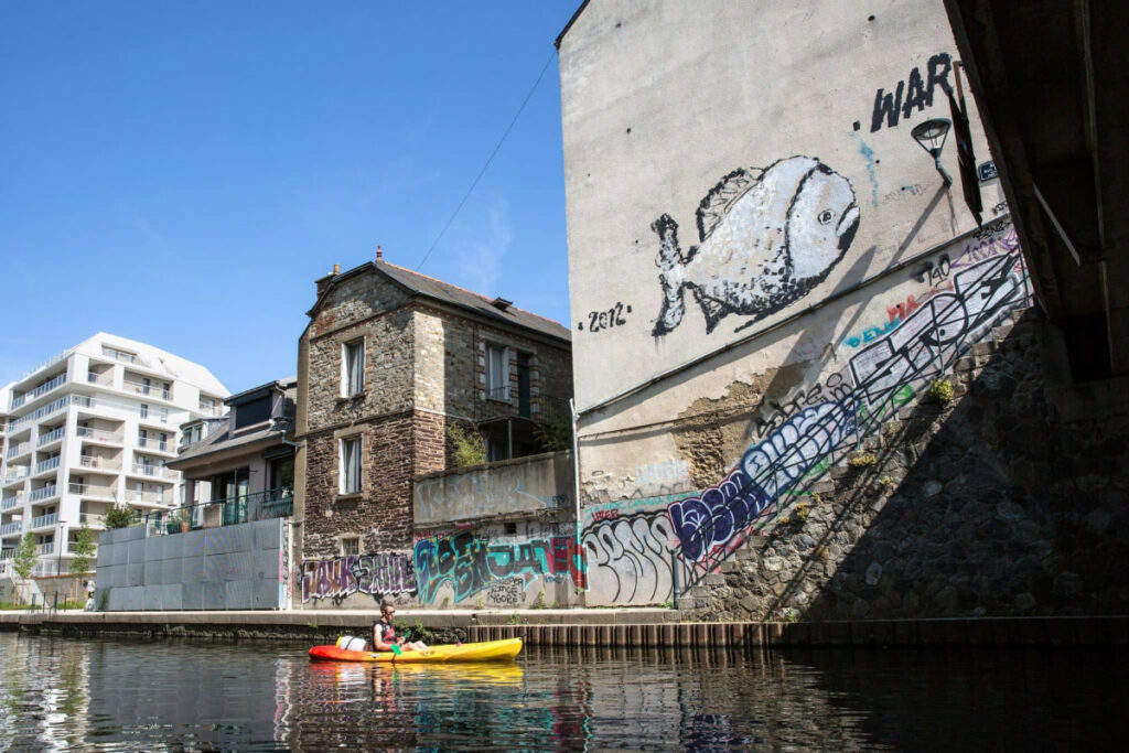 Canoe navigant sur la Vilaine. Du street art est visible sur les murs des bâtiments.